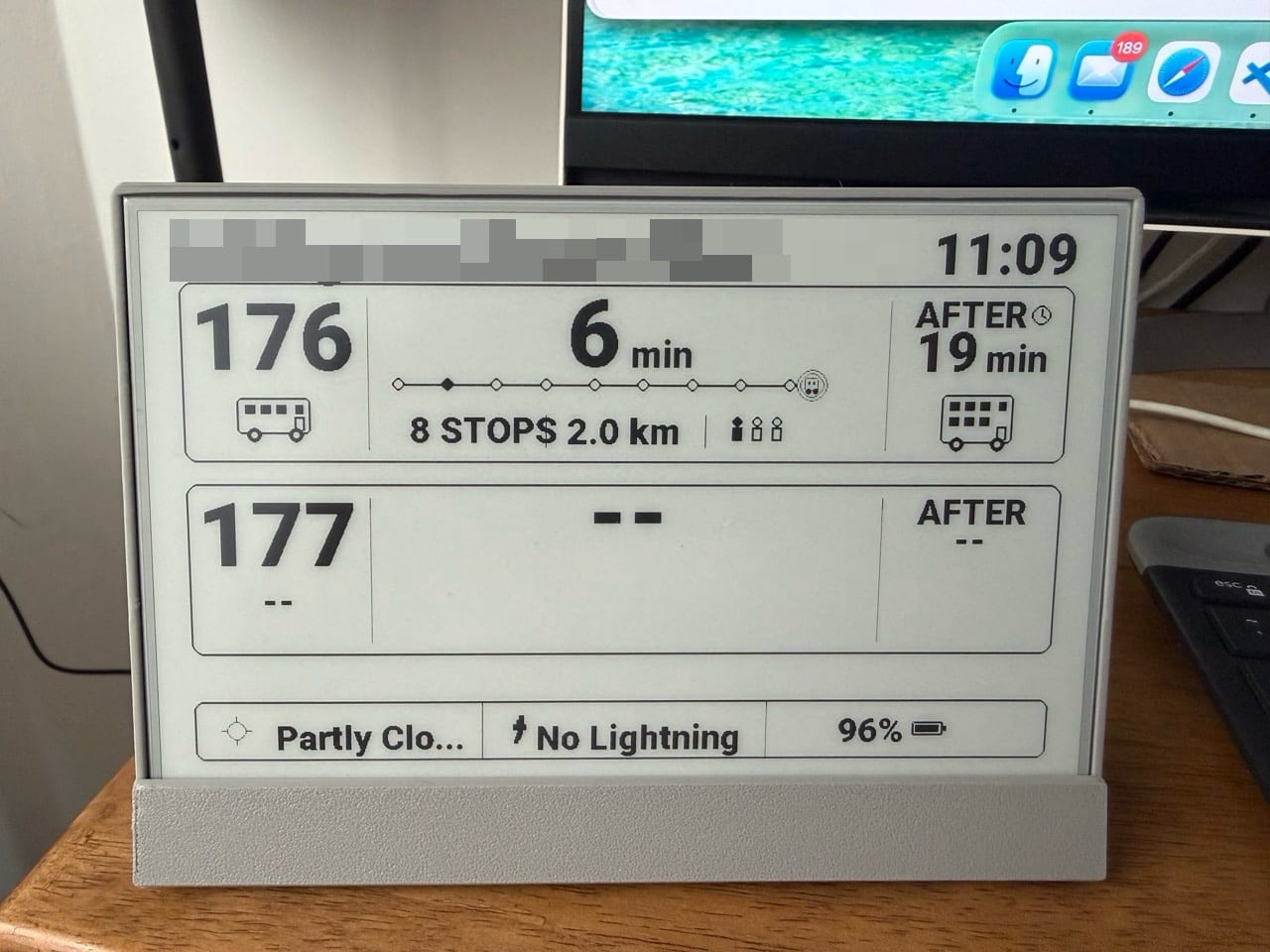 The finished e-paper screen in a grey stand on a desk, showing service 176 arriving in 6 minutes with a row of stops and an occupancy icon, and a footer with weather, lightning and battery level